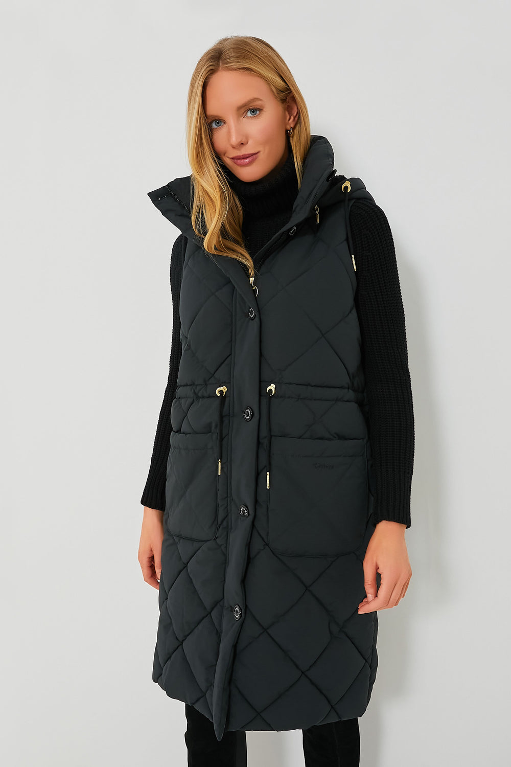 A woman wears a long, black, quilted Barbour sleeveless vest with a removable sherpa hood over a black turtleneck sweater.