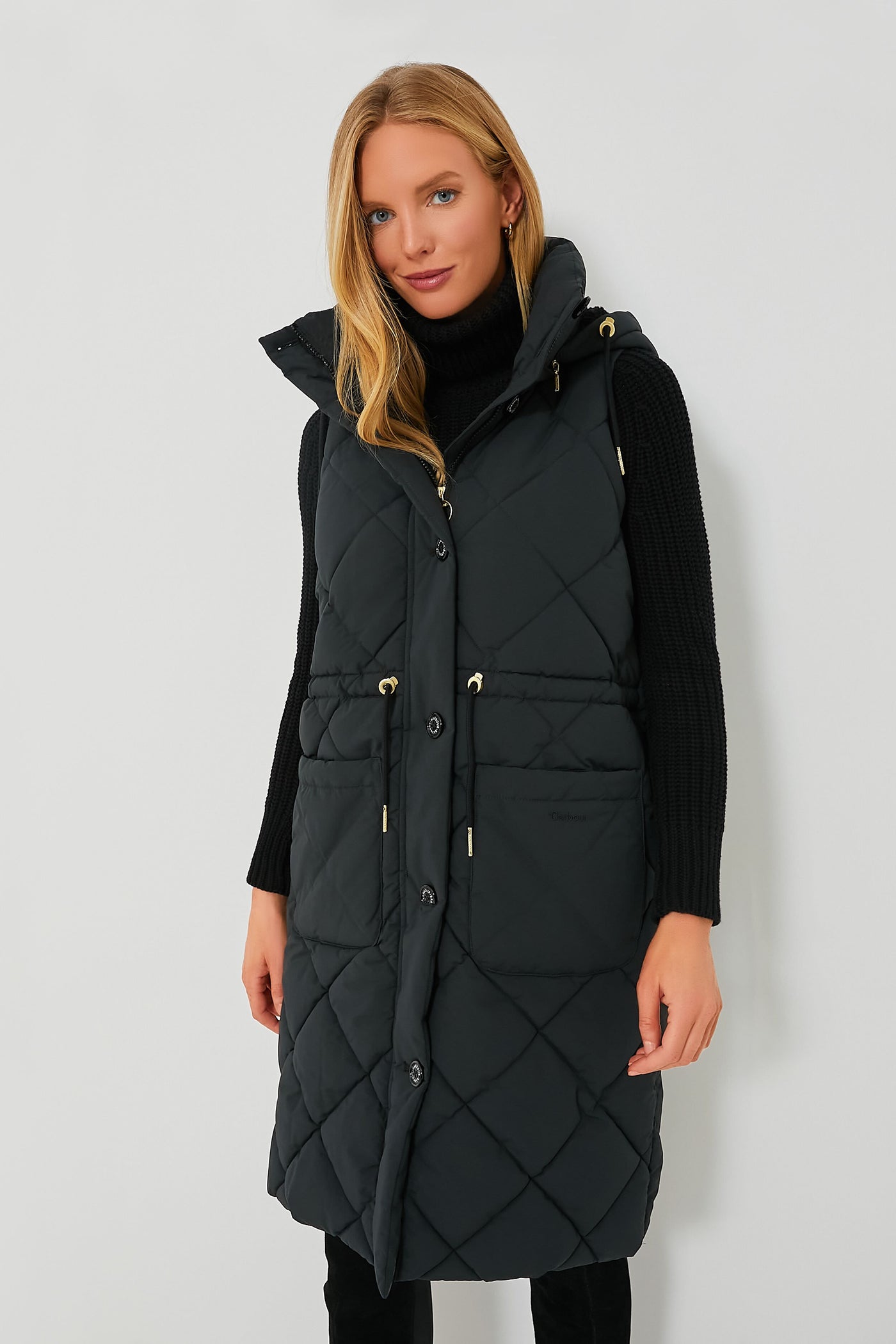 A woman wears a long, black, quilted Barbour sleeveless vest with a removable sherpa hood over a black turtleneck sweater.