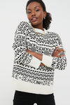 A woman poses with folded arms, wearing a Tuckernuck black and white Fair Isle sweater with "SAISONNAIRE" on the front and black pants.