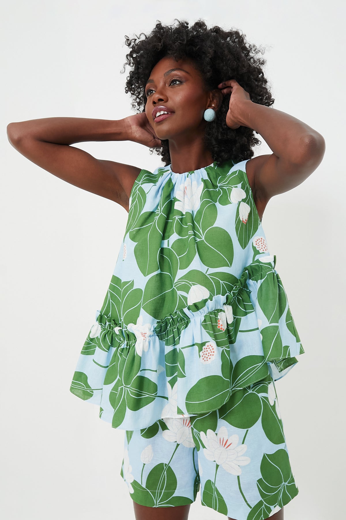 A woman in a Borgo de Nor Green Waterlily Alora Top with an asymmetric ruffle hem and matching green and white floral shorts stands with her hands touching her hair.
