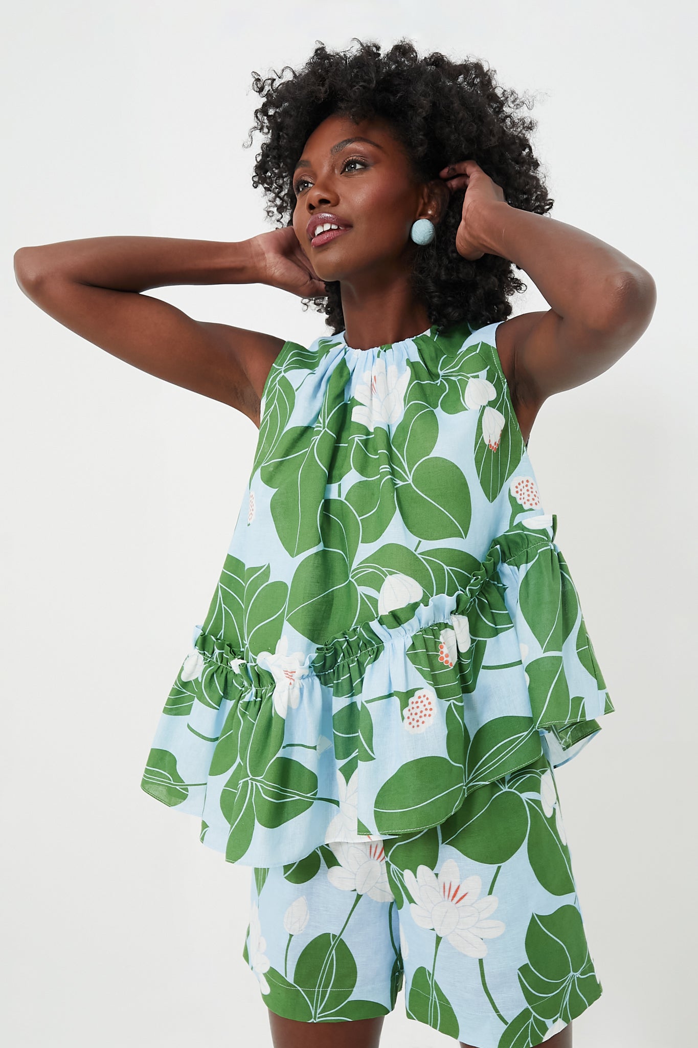 A woman in a Borgo de Nor Green Waterlily Alora Top with an asymmetric ruffle hem and matching green and white floral shorts stands with her hands touching her hair.