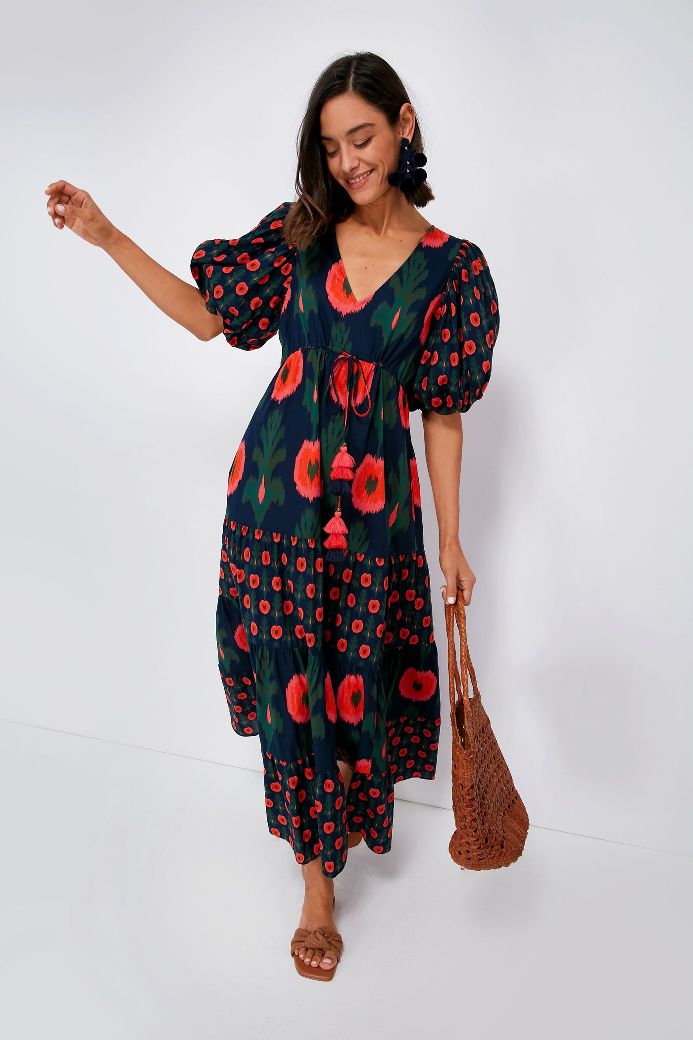 A woman wearing a Figue navy blue bohemian maxi dress with red floral print and puff sleeves, paired with a brown woven bag—ideal for luxury resort wear.