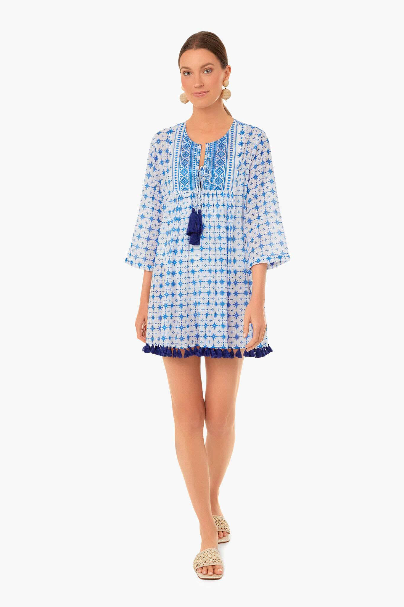A woman wearing a vacation-ready blue and white Ro's Garden tunic dress with tassels, large round earrings, and woven slide sandals.