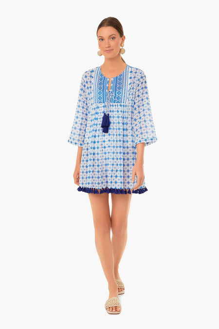 A woman wearing a vacation-ready blue and white Ro's Garden tunic dress with tassels, large round earrings, and woven slide sandals.