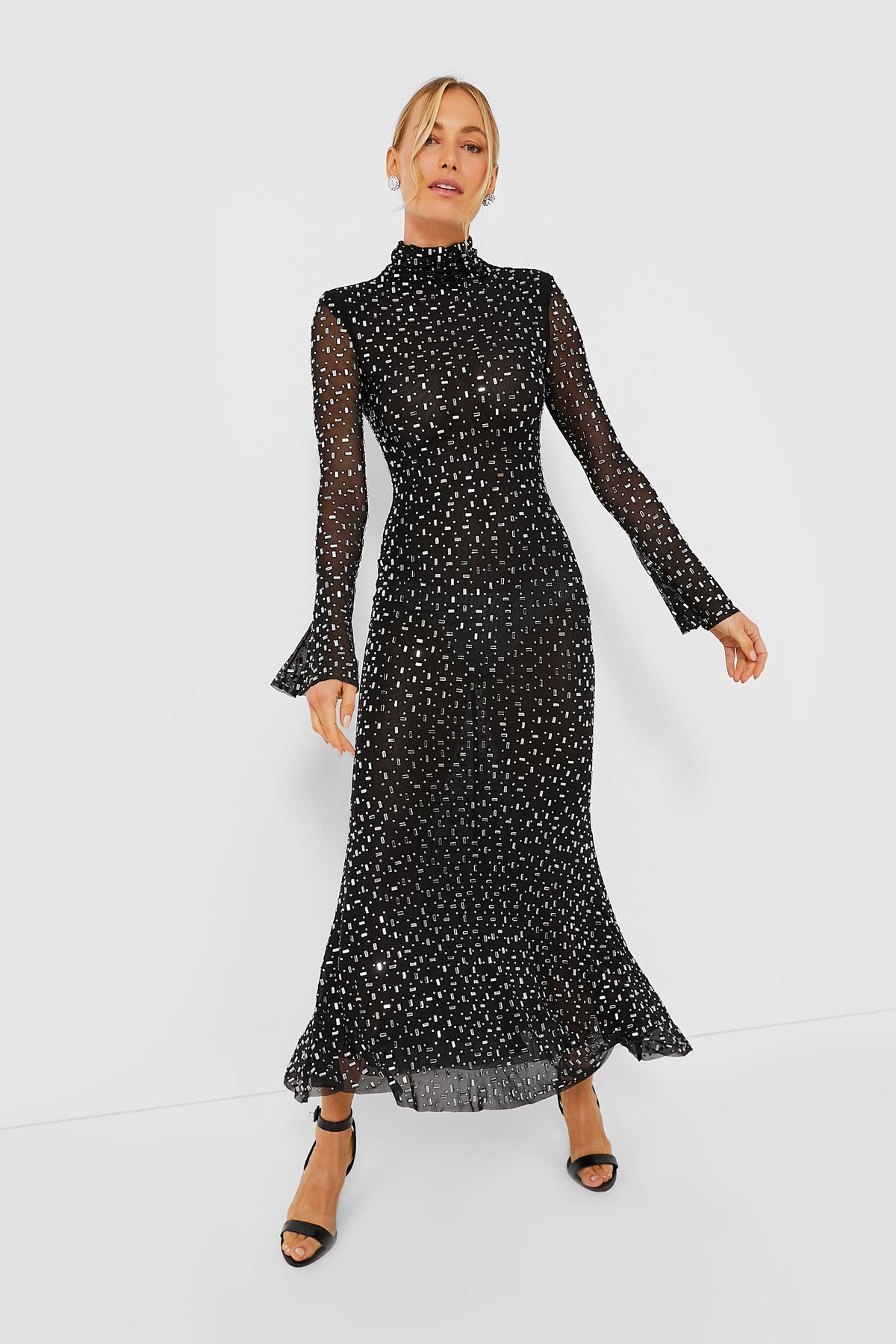 A woman wearing a Self-Portrait black square rhinestone mesh dress with sheer long sleeves, white polka dots, high neck, and ruffled cuffs and hem, styled with black open-toe heels.