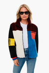 A woman in KULE sunglasses, white t-shirt, blue jeans, and a cozy color-block fleece jacket—featuring red, blue, yellow, white, and black sections—stands confidently in a boxy fit.