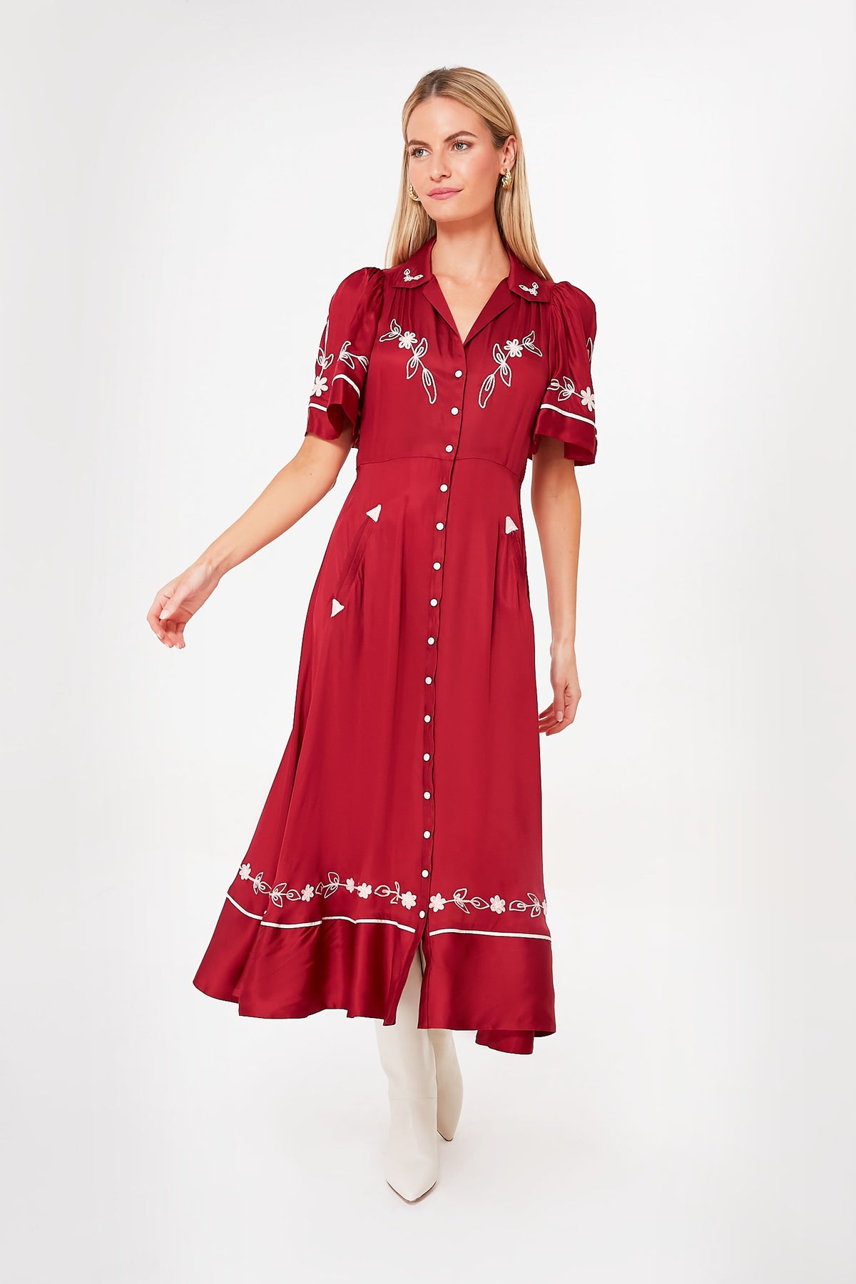 A woman wears THE GREAT.'s Rhubarb Bridge Dress, a vintage red midi dress with white floral embroidery and short puff sleeves, paired with white boots.