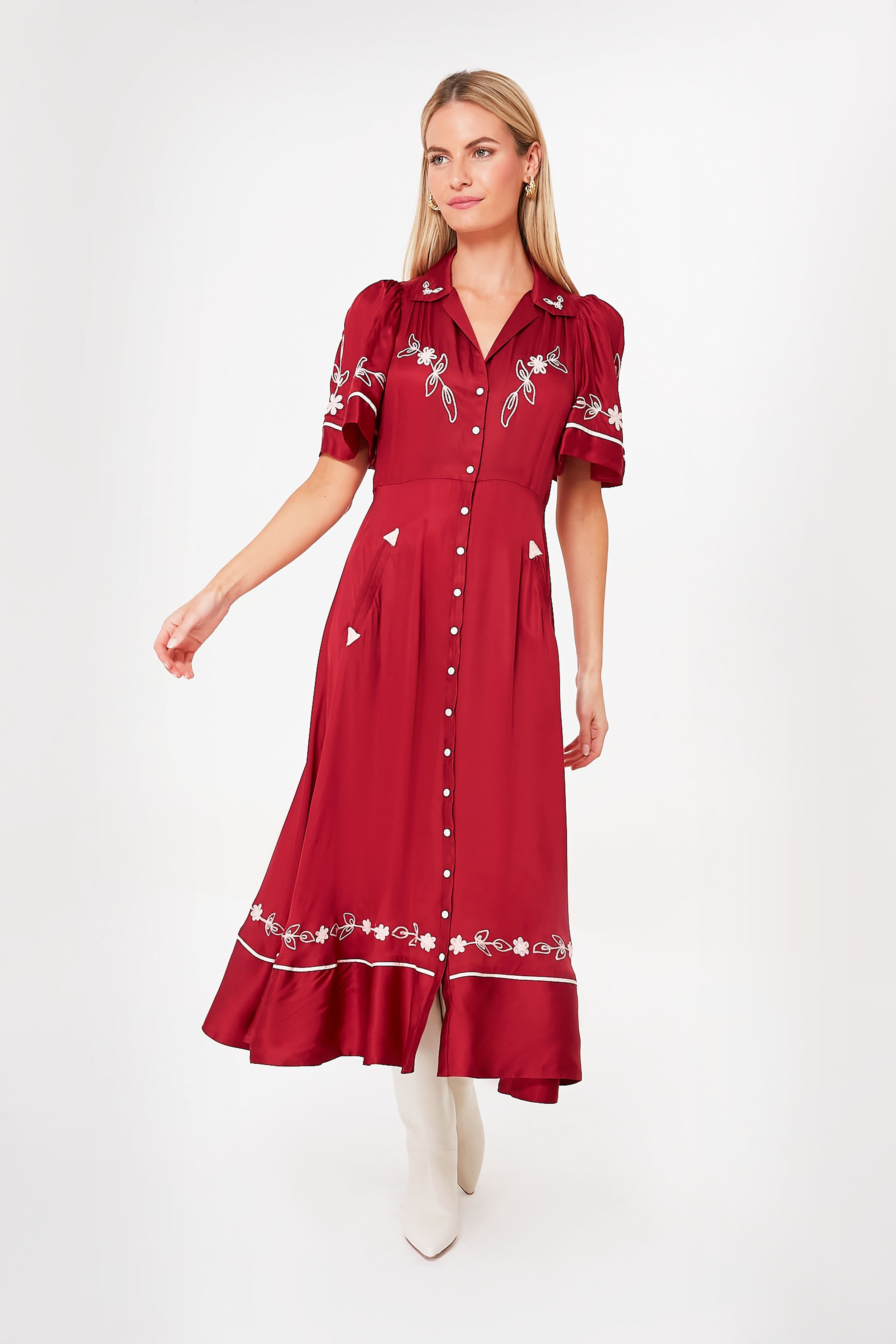 A woman wears THE GREAT.'s Rhubarb Bridge Dress, a vintage red midi dress with white floral embroidery and short puff sleeves, paired with white boots.