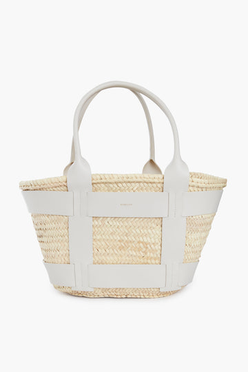 The Demellier woven straw luxury tote features two white leather handles and side accents, highlighting the elegance of handcrafted handbags.
