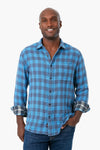 A person in Faherty's blue plaid organic cotton button-up shirt with rolled sleeves and blue jeans, smiling brightly.