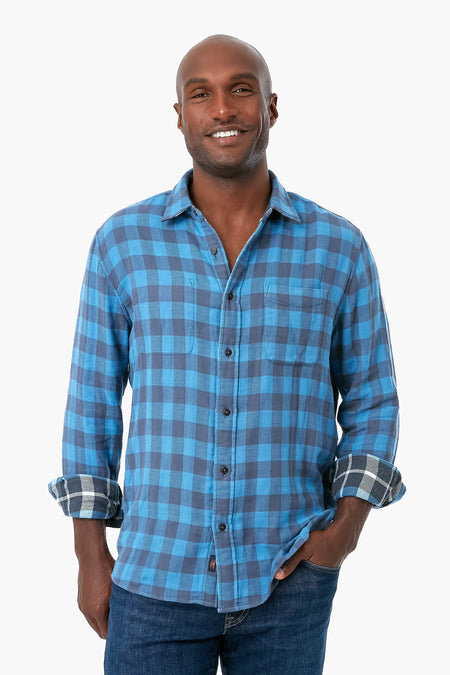 A person in Faherty's blue plaid organic cotton button-up shirt with rolled sleeves and blue jeans, smiling brightly.