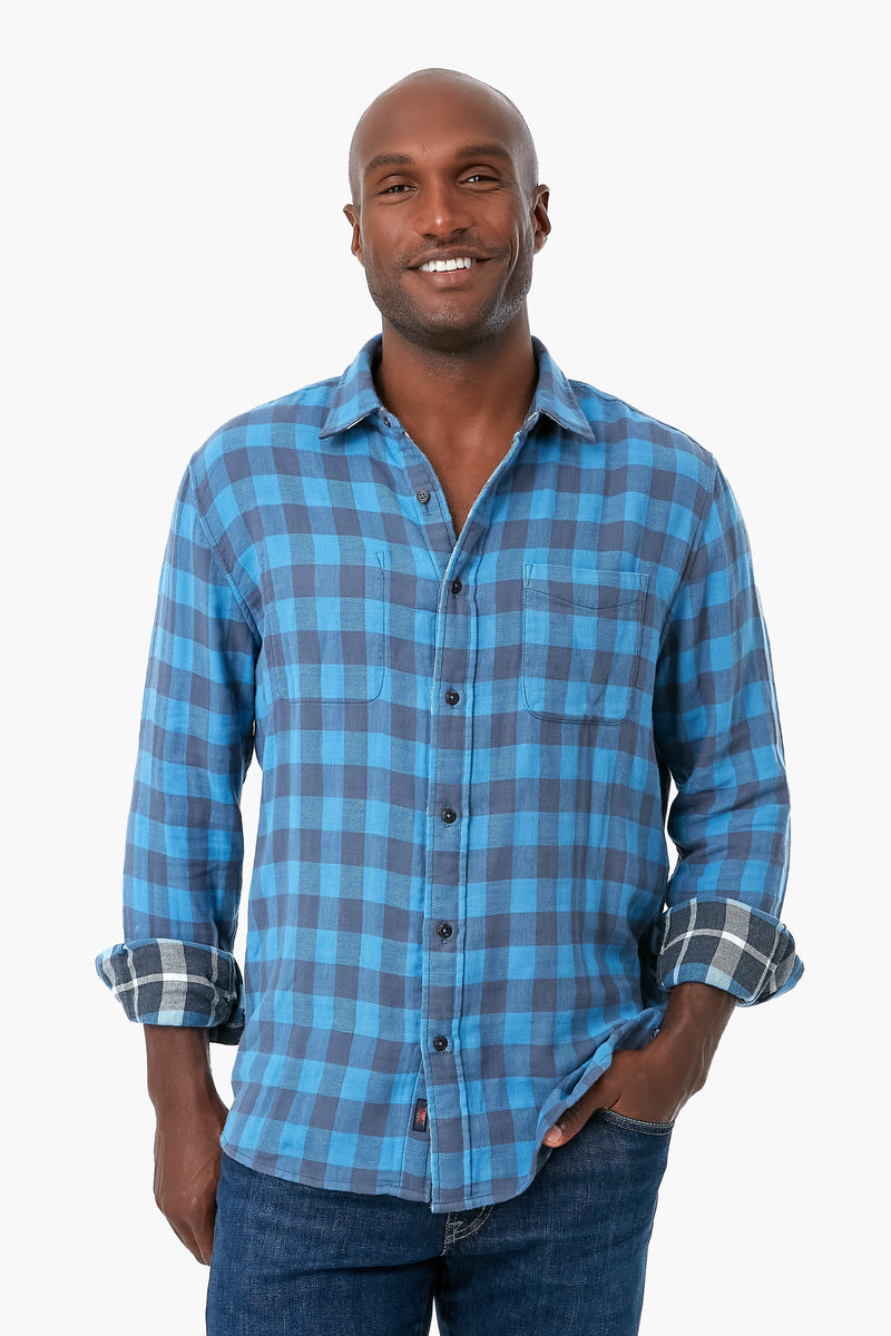 A person in Faherty's blue plaid organic cotton button-up shirt with rolled sleeves and blue jeans, smiling brightly.