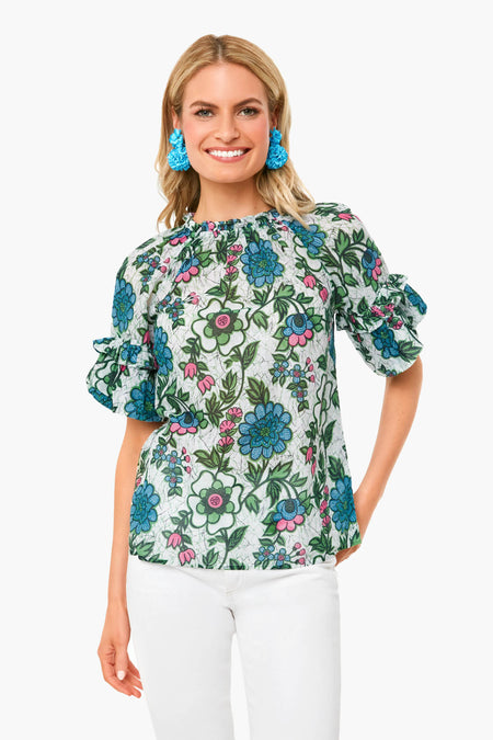 A woman stands smiling in a Banjanan white floral top with blue and green patterns made from organic cotton, paired with white pants and blue tassel earrings.