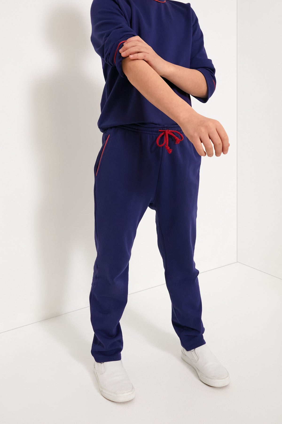 A person stands in a corner wearing Oso & Me’s navy blue tracksuit with red trim and white slip-on shoes. The stylish kids jogger pants are highlighted, with only the lower face and body visible.