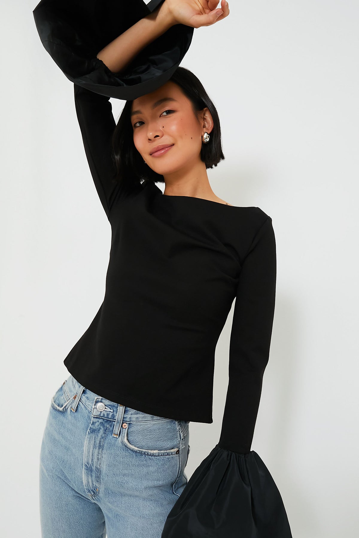 A woman poses in a STAUD Black Paige Top with a bateau neckline and large, flared cuffs, styled with light blue jeans.