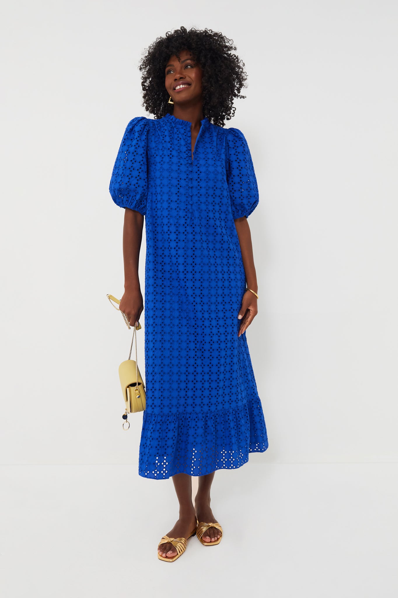 A woman wears a bright blue Hyacinth House Tryall Dress with puff sleeves and eyelet fabric, gold sandals, and carries a small beige handbag.