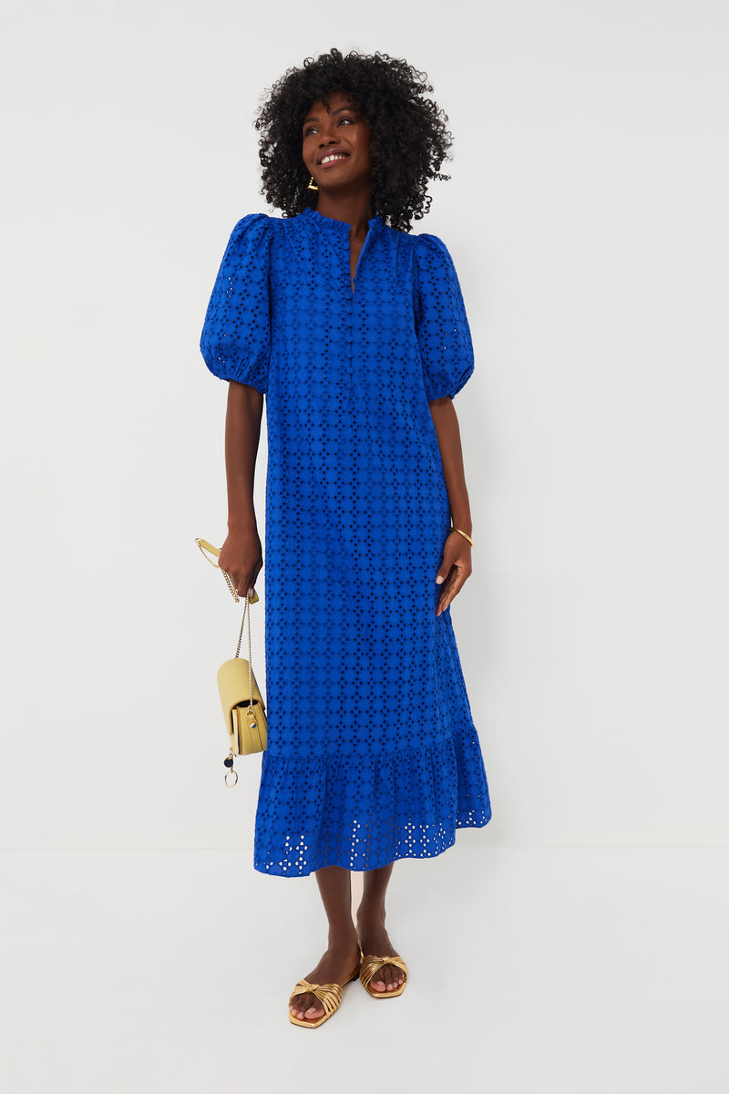 A woman wears a bright blue Hyacinth House Tryall Dress with puff sleeves and eyelet fabric, gold sandals, and carries a small beige handbag.
