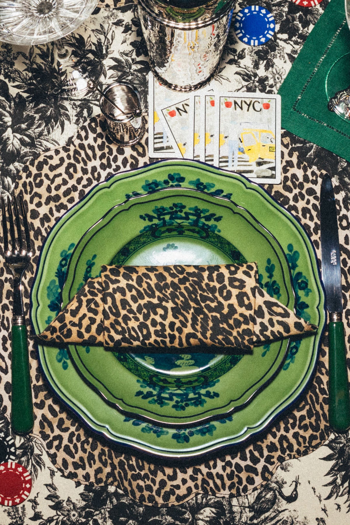A green Ginori 1735 Oriente Italiano Malachite porcelain dinner plate with a leopard print napkin is set on a patterned tablecloth, alongside silverware, playing cards, sunglasses, and poker chips.