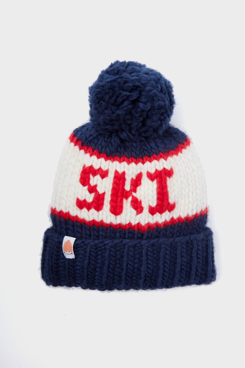The Sh*t That I Knit beanie is handcrafted from soft Peruvian Merino wool, with a blue pom-pom, blue brim, and SKI in bold red letters with red accents.