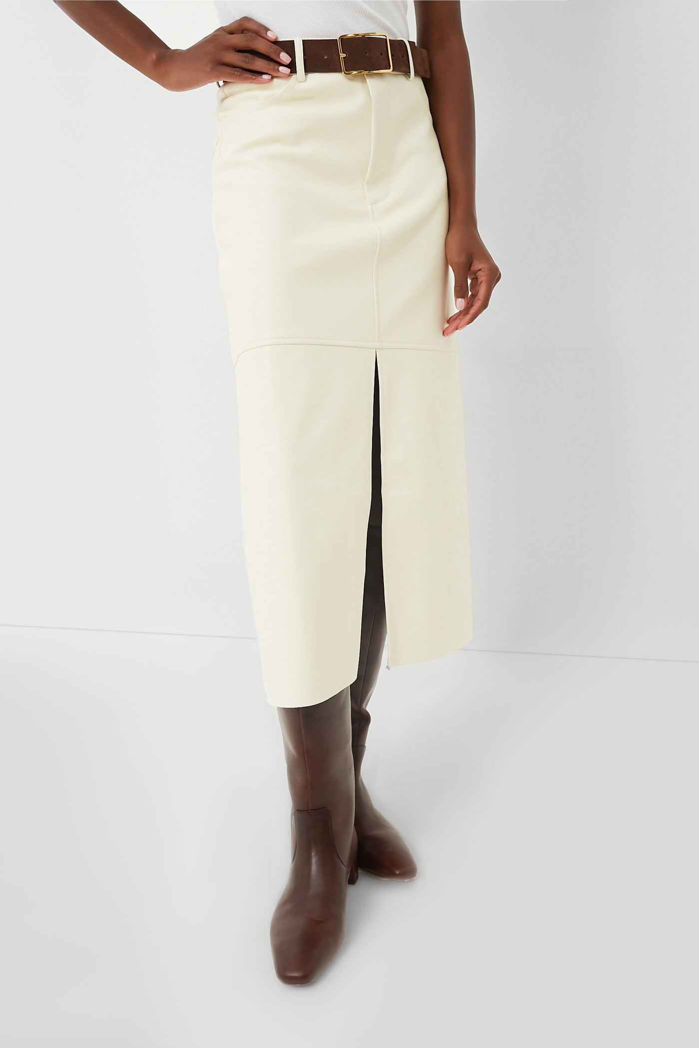 A person wears a WeWoreWhat ecru vegan leather high-rise midi skirt with a front slit, styled with a brown belt and knee-high brown boots.