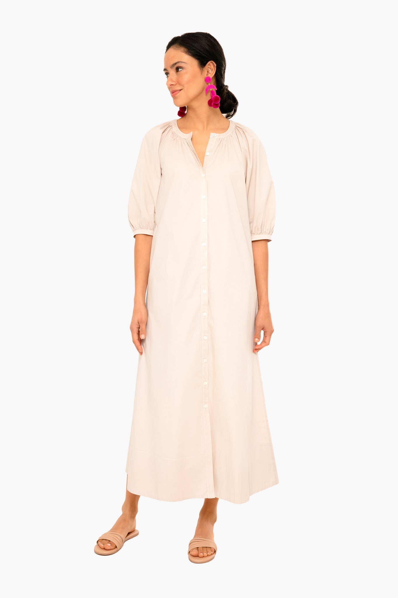 A woman wears the STAUD Vincent Dress—a light beige, button-down caftan with three-quarter sleeves—styled with pink statement earrings and flat sandals for an effortless look.