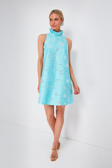 A woman wears a Tuckernuck Blythe dress—a sleeveless light blue floral fil coupe mini dress with a ruffle stand collar—paired with gold open-toe heels.