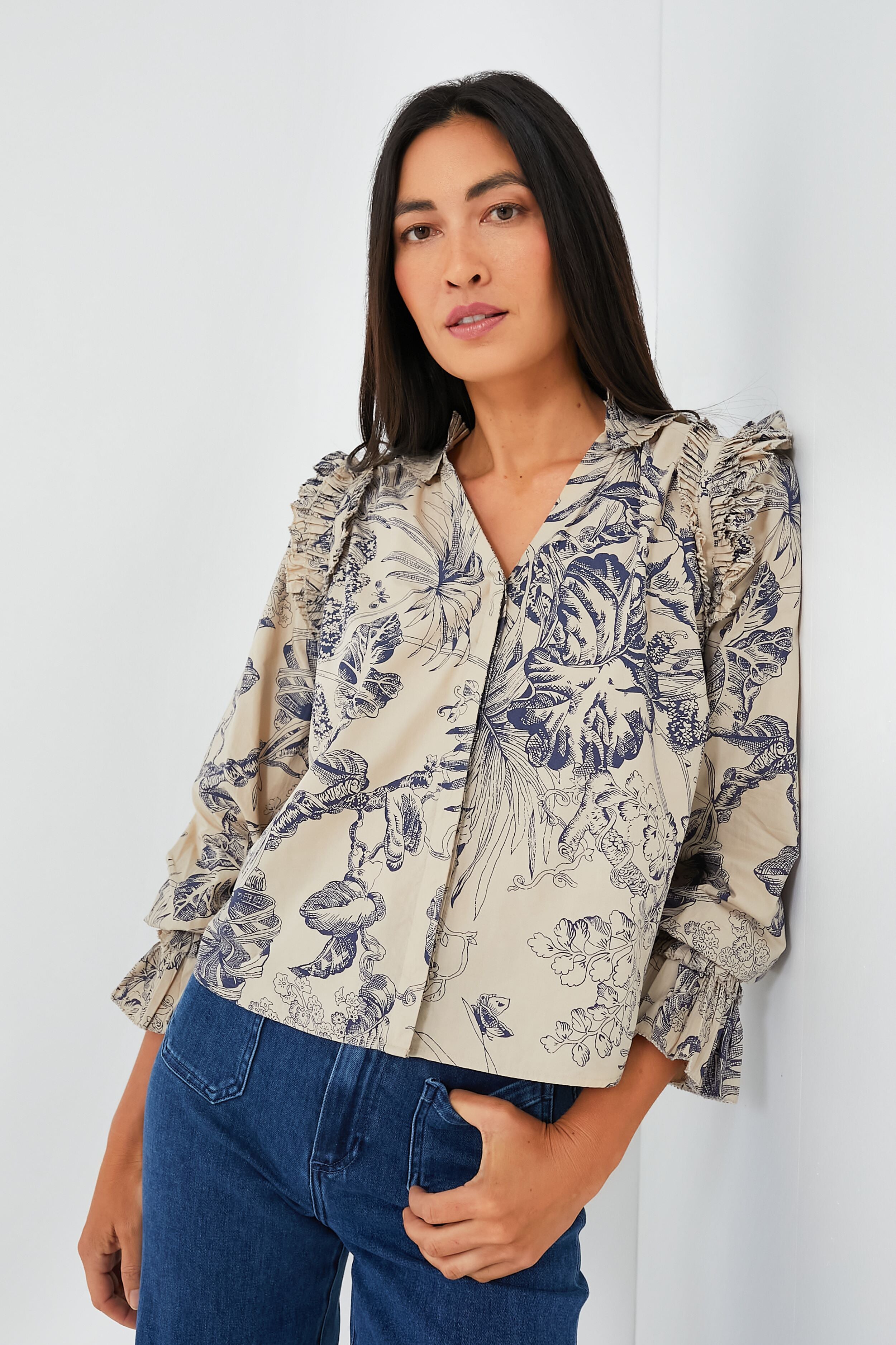 A woman with long dark hair wears a Love the Label Jessie Top, a beige blouse with dark floral print and ruffled details, paired with blue jeans.
