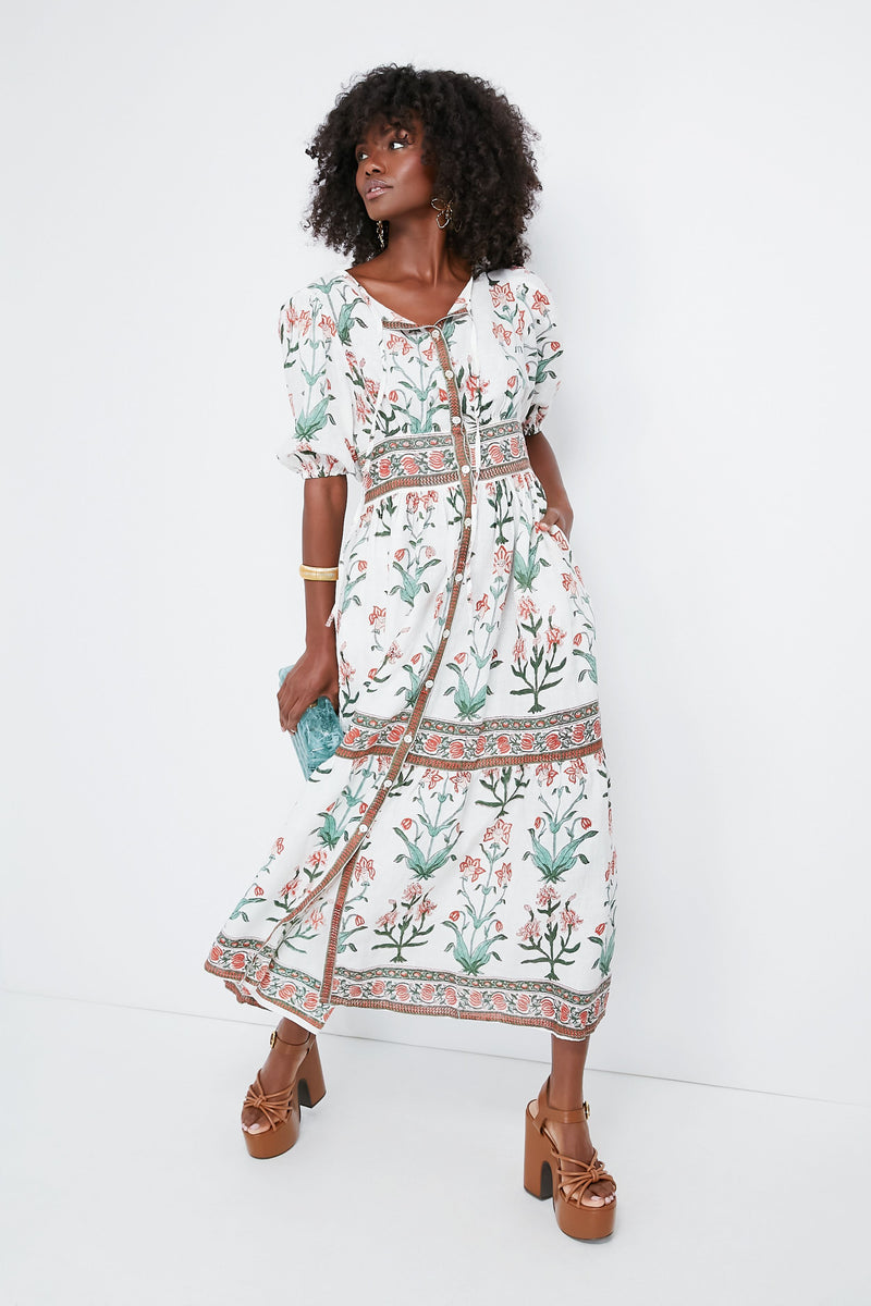 A woman wears a white floral print boho maxi dress with puff sleeves by Hannah Artwear, holding a blue clutch and wearing brown platform sandals.
