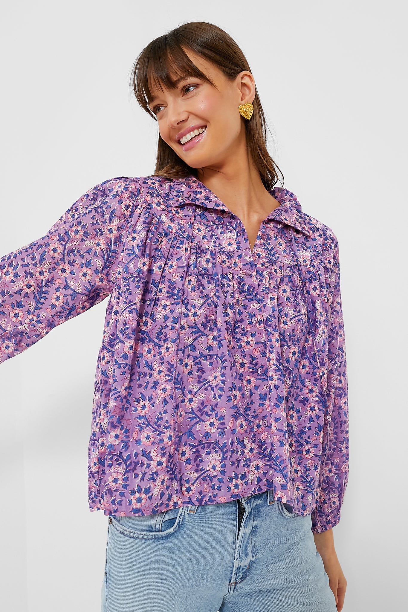 A woman in a violet wildflower blouse from Love Tomas by Emerson Fry and light blue jeans smiles, gazing to the side.