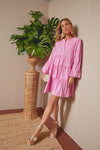 A woman in a pink, long-sleeve, tiered floral block print dress by Juliet Dunn stands beside a woven plant stand with lush green leaves. She completes the luxury resort-ready look with platform sandals and statement earrings.