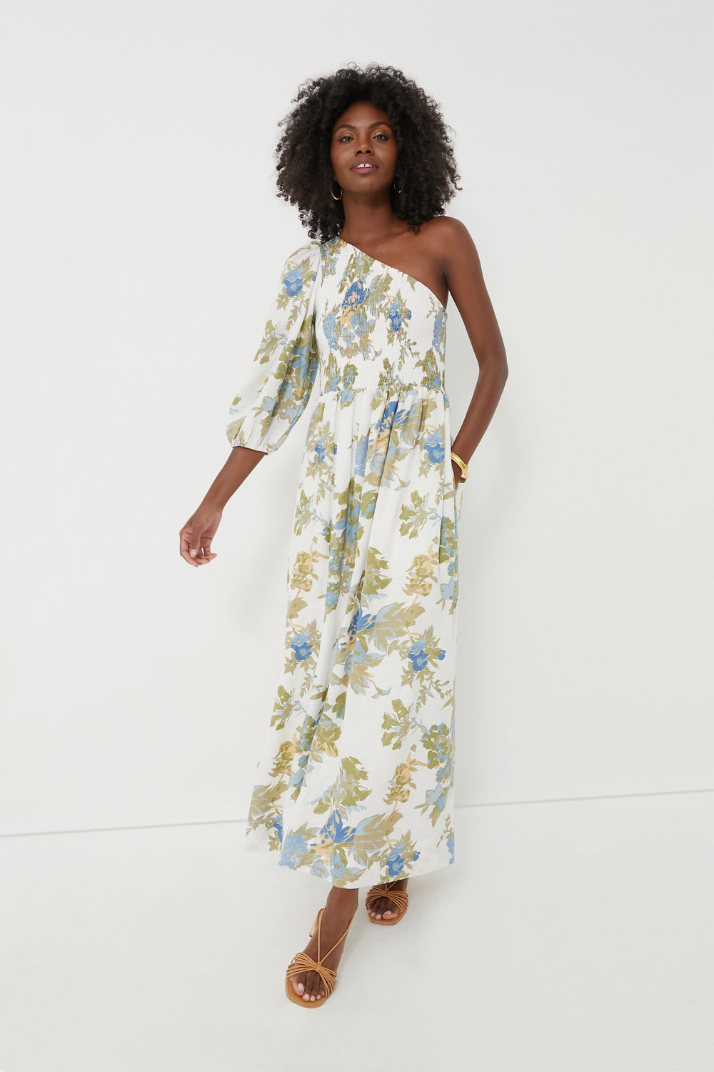 A woman wears the FAITHFULL THE BRAND Ivory Escala Floral Anha Dress, a white one-shoulder handmade Bali maxi dress with green and blue prints.