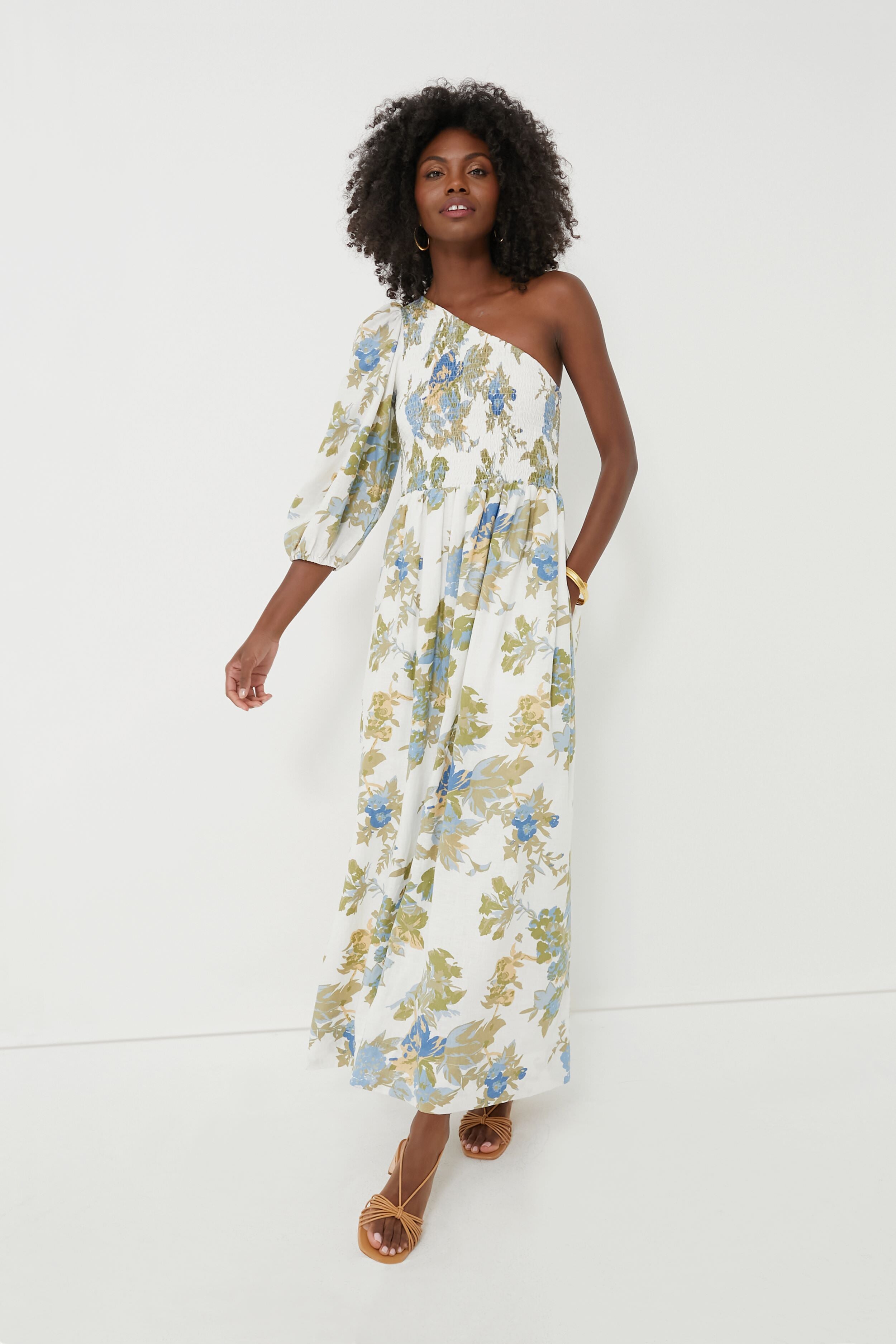 A woman wears the FAITHFULL THE BRAND Ivory Escala Floral Anha Dress, a white one-shoulder handmade Bali maxi dress with green and blue prints.