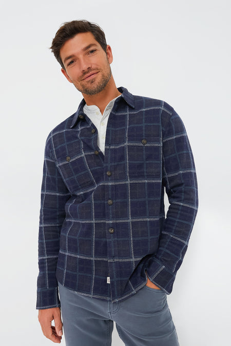 A man wearing a Marine Layer navy blue plaid button-up shirt made from eco-friendly materials over a white top and gray pants.