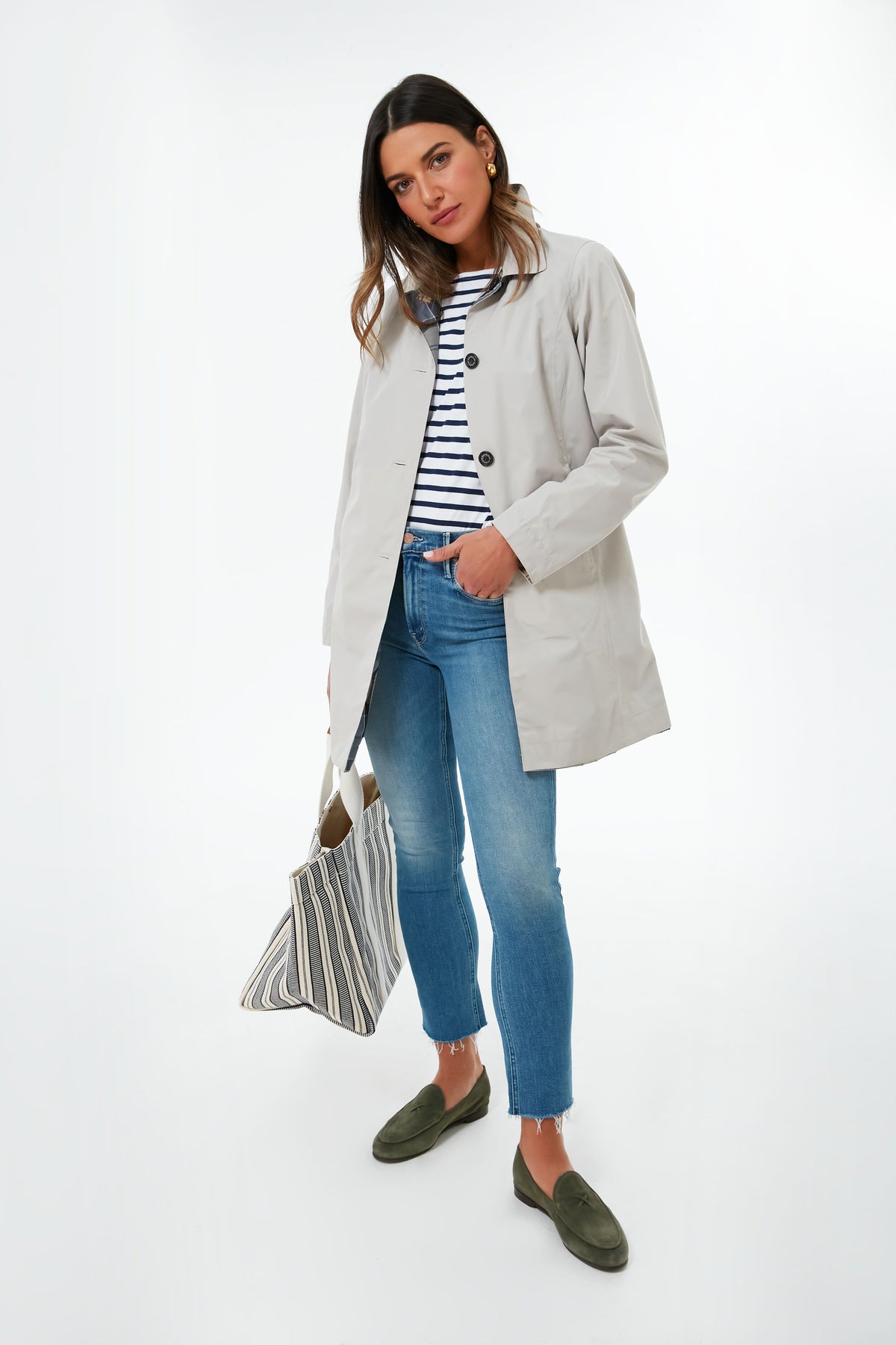 A woman wears a light beige Barbour raincoat over a striped shirt, blue jeans, and green loafers, and carries a striped tote bag.