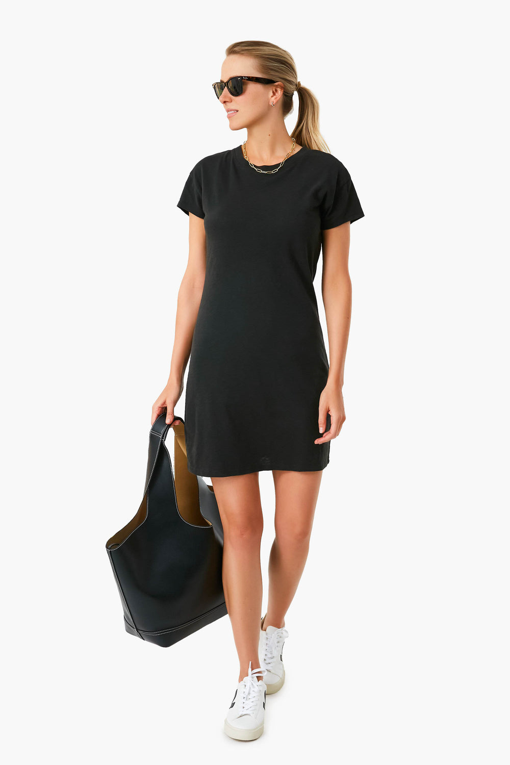 A woman in a Z SUPPLY Black Modern Slub Tee Dress, paired with white sneakers and sunglasses, carries a large black tote bag.