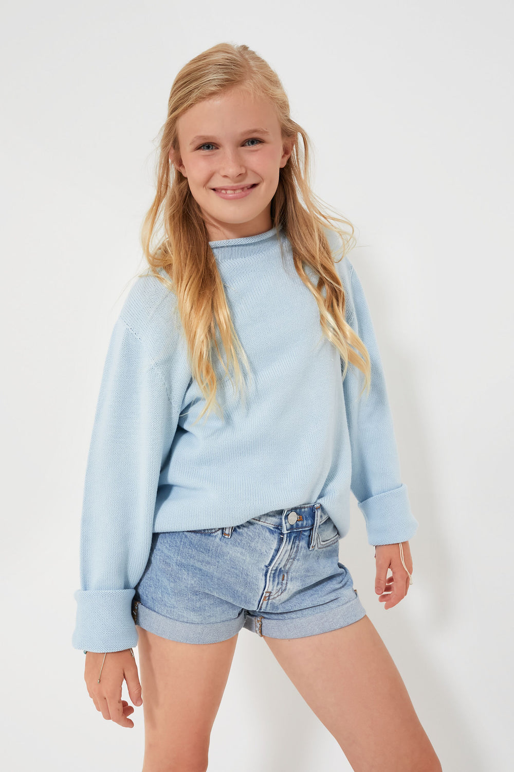A girl smiles, wearing a light blue kids cotton sweater by A Soft Idea and denim shorts.