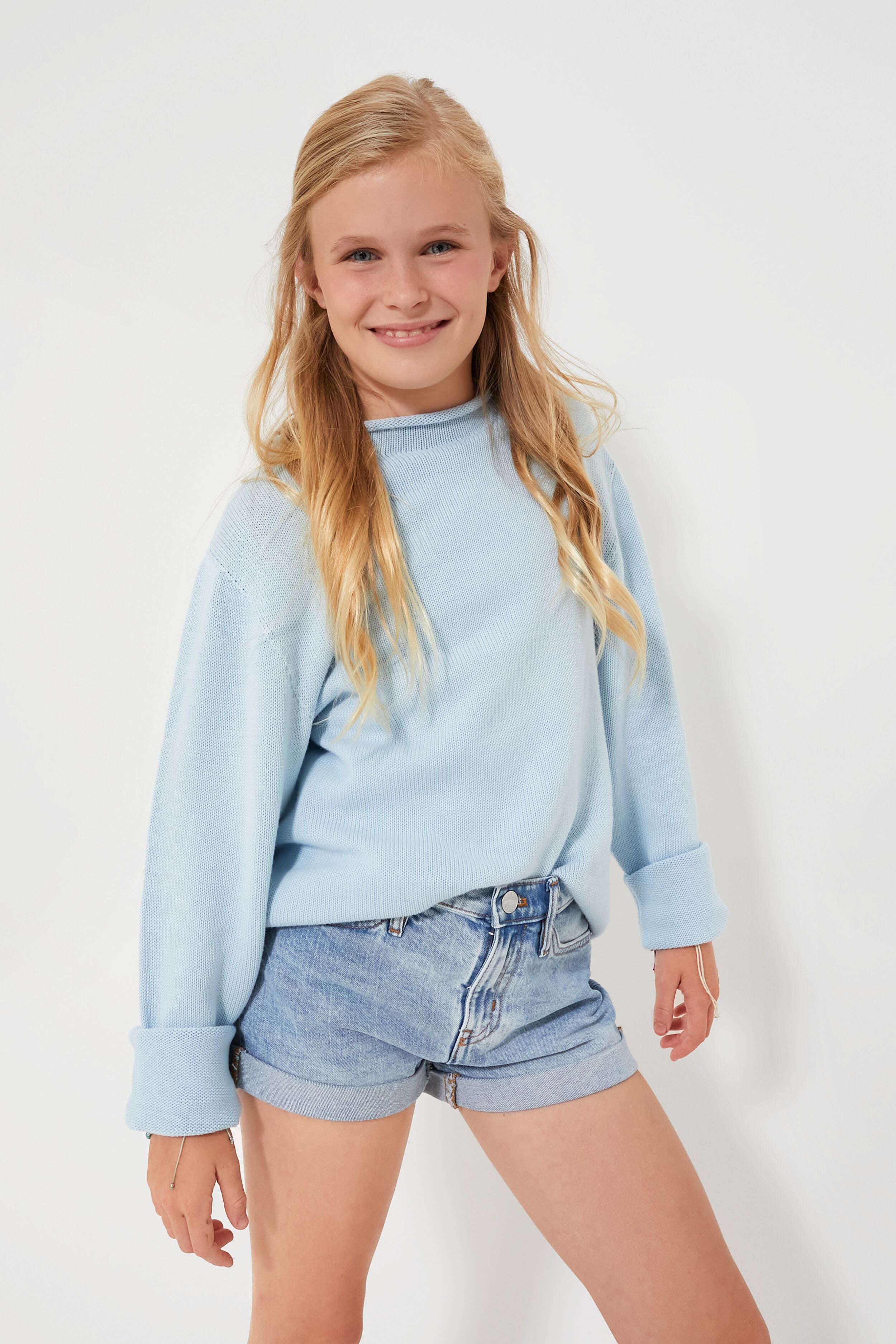 A girl smiles, wearing a light blue kids cotton sweater by A Soft Idea and denim shorts.