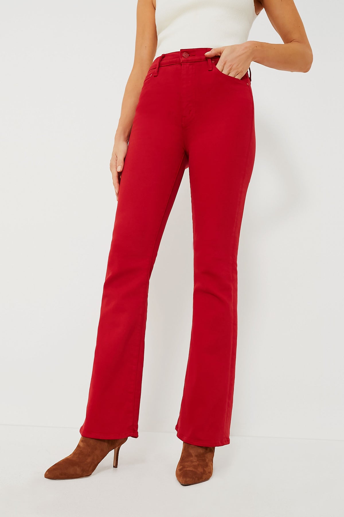 A person stands confidently in high-rise flared red denim jeans from MOTHER, paired with a white sleeveless top and brown heeled boots.