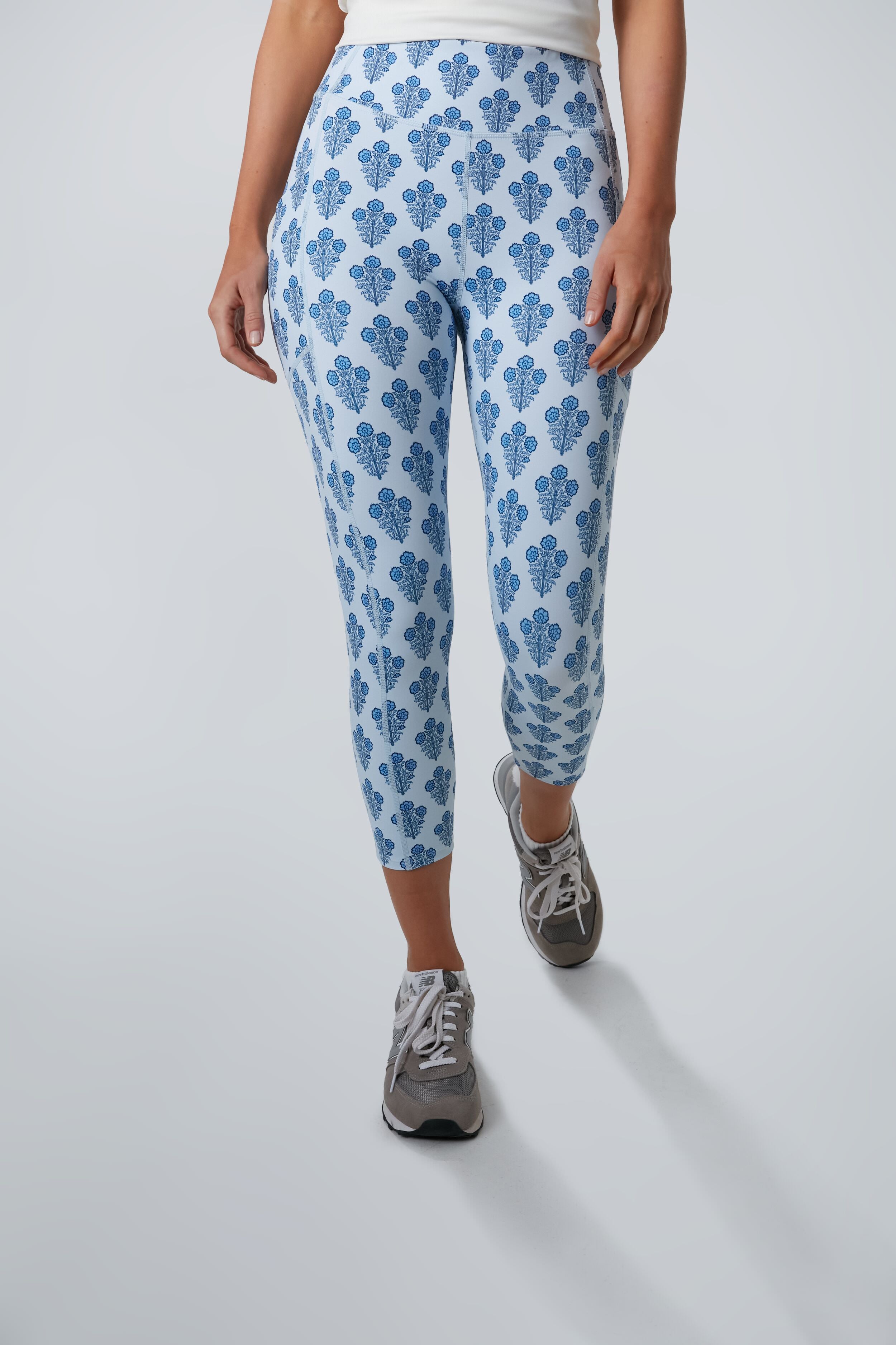 A person wearing Tnuck Sport light blue floral leggings, a Kaylee Capri white top, and gray sneakers stands in a well-lit space.