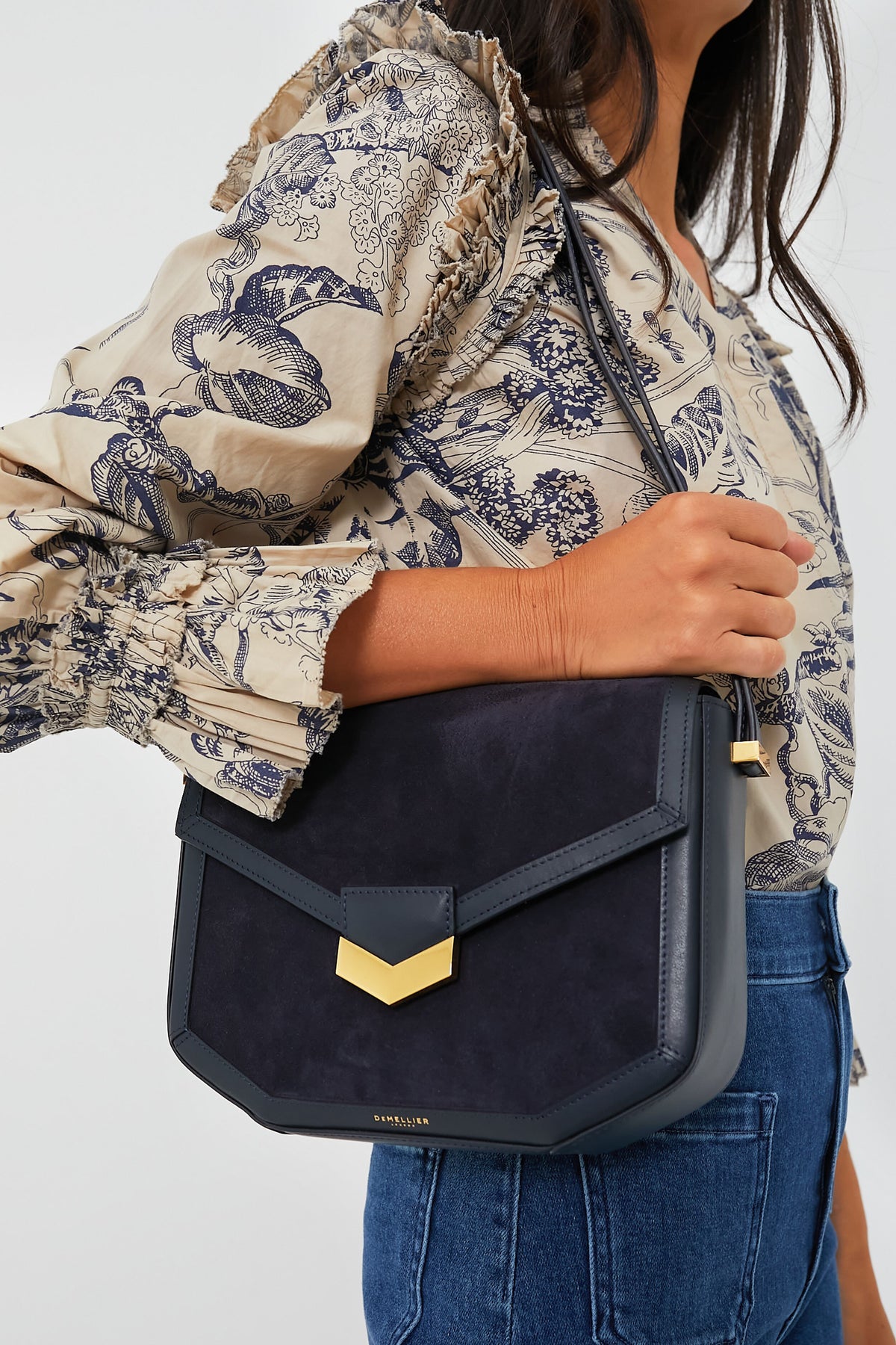 A woman in a patterned blouse and jeans carries a Demellier black and gold crossbody bag.