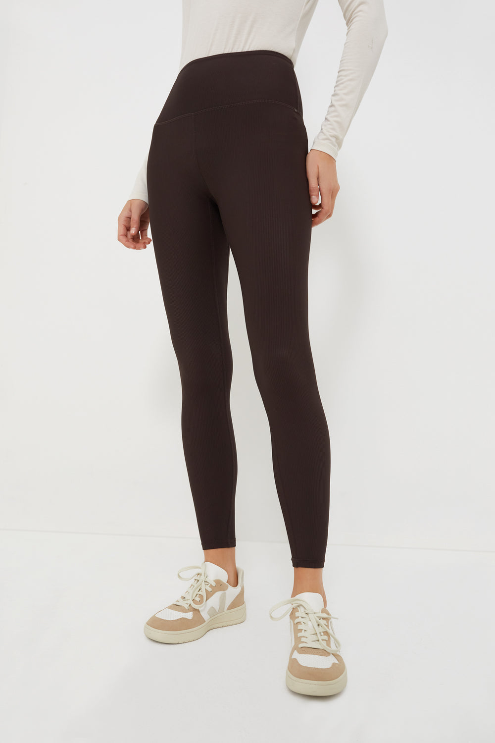 A person wears a long-sleeve beige Varley top, black high-rise athleisure leggings by Varley, and beige sneakers.