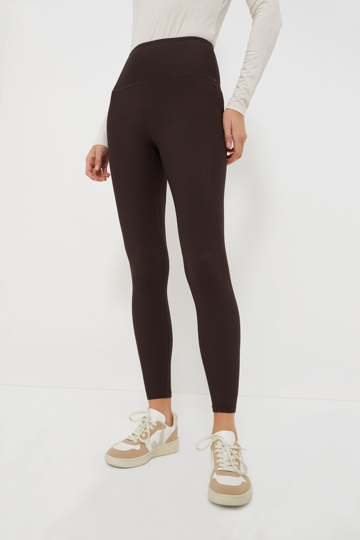 A person wears a long-sleeve beige Varley top, black high-rise athleisure leggings by Varley, and beige sneakers.