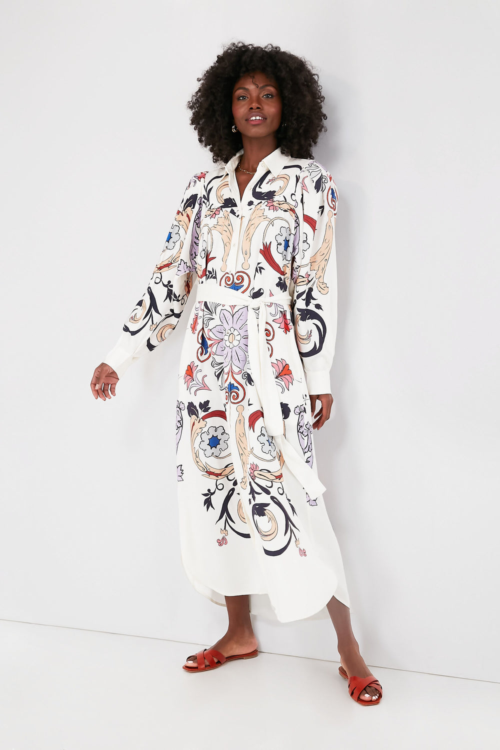 A woman wears a Carolina K. Luna Dress, a long-sleeve white shirtdress with colorful abstract designs, styled with red flat sandals.