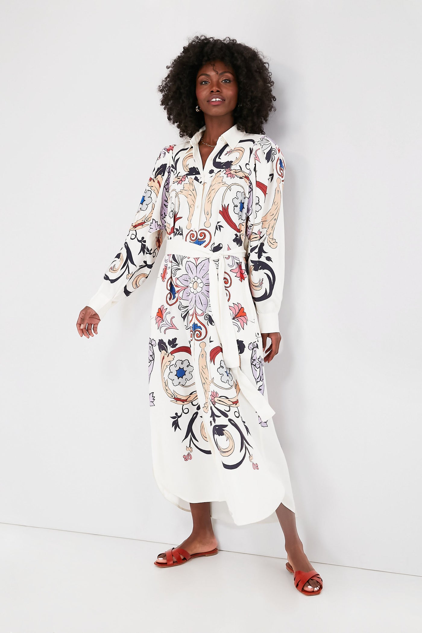 A woman wears a Carolina K. Luna Dress, a long-sleeve white shirtdress with colorful abstract designs, styled with red flat sandals.