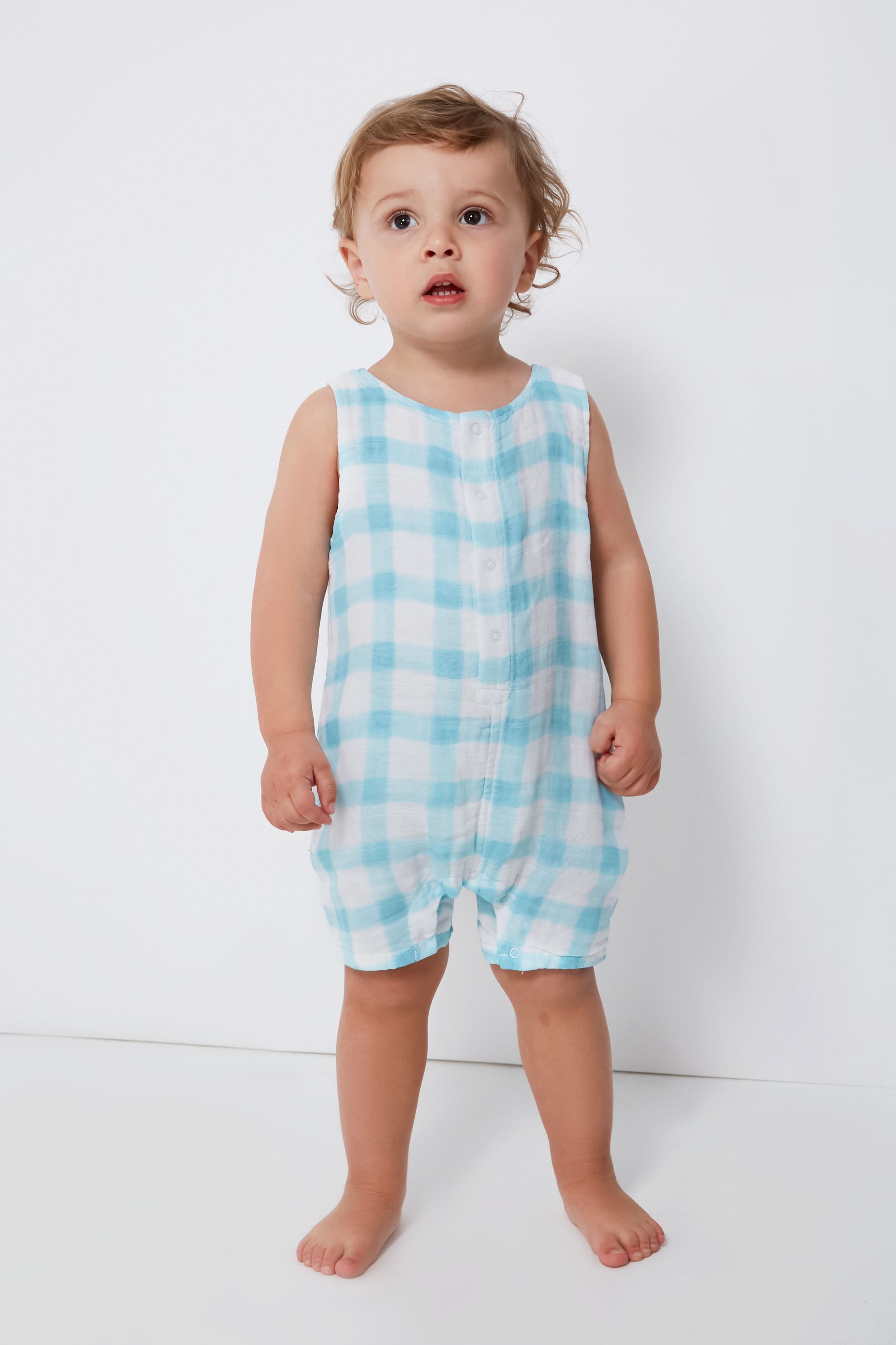 A toddler stands barefoot, dressed in an Angel Dear sleeveless blue and white plaid baby romper crafted from soft organic cotton.