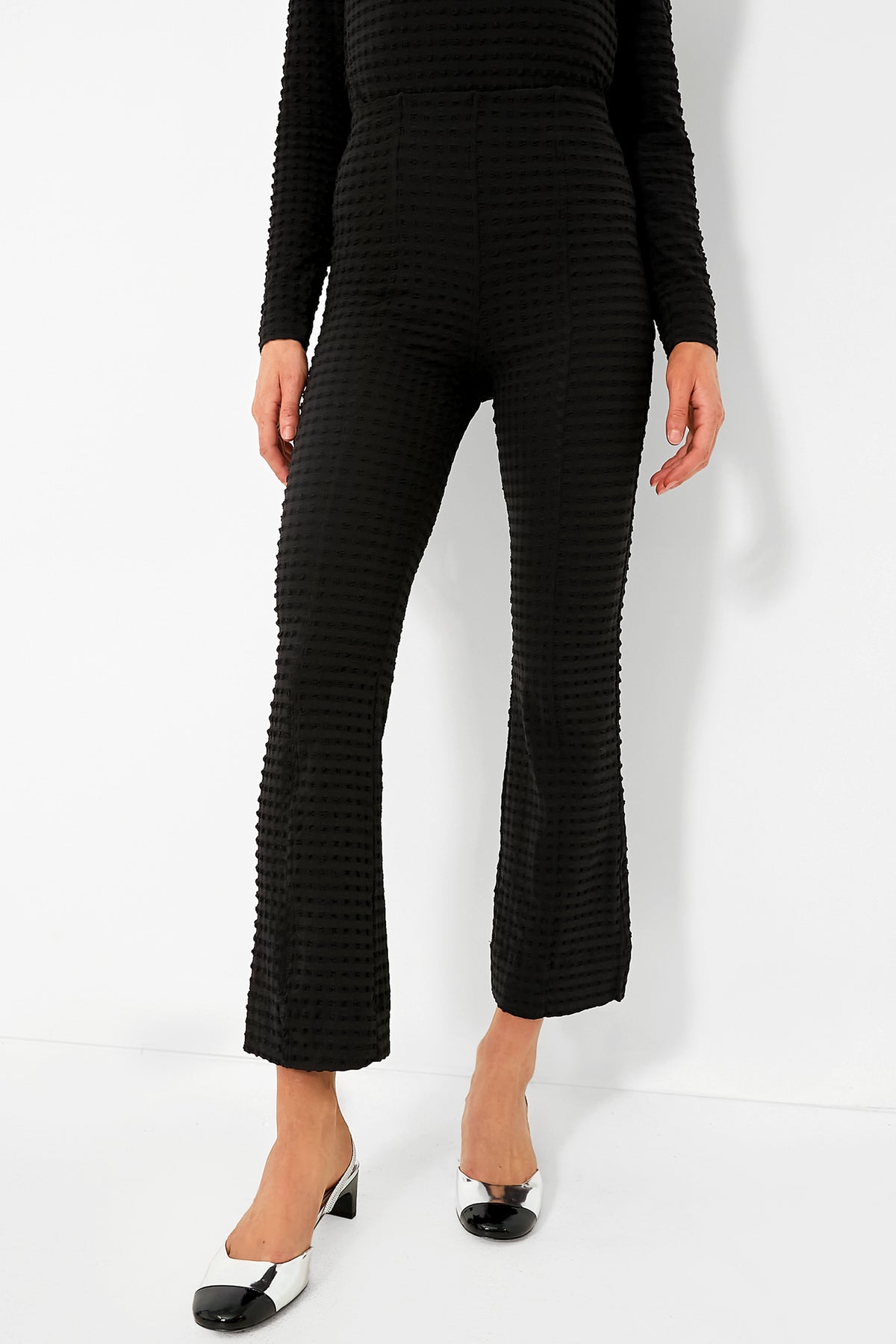 A person wears black textured GANNI pants with a matching long-sleeve top, styled with black and white low-heeled shoes.