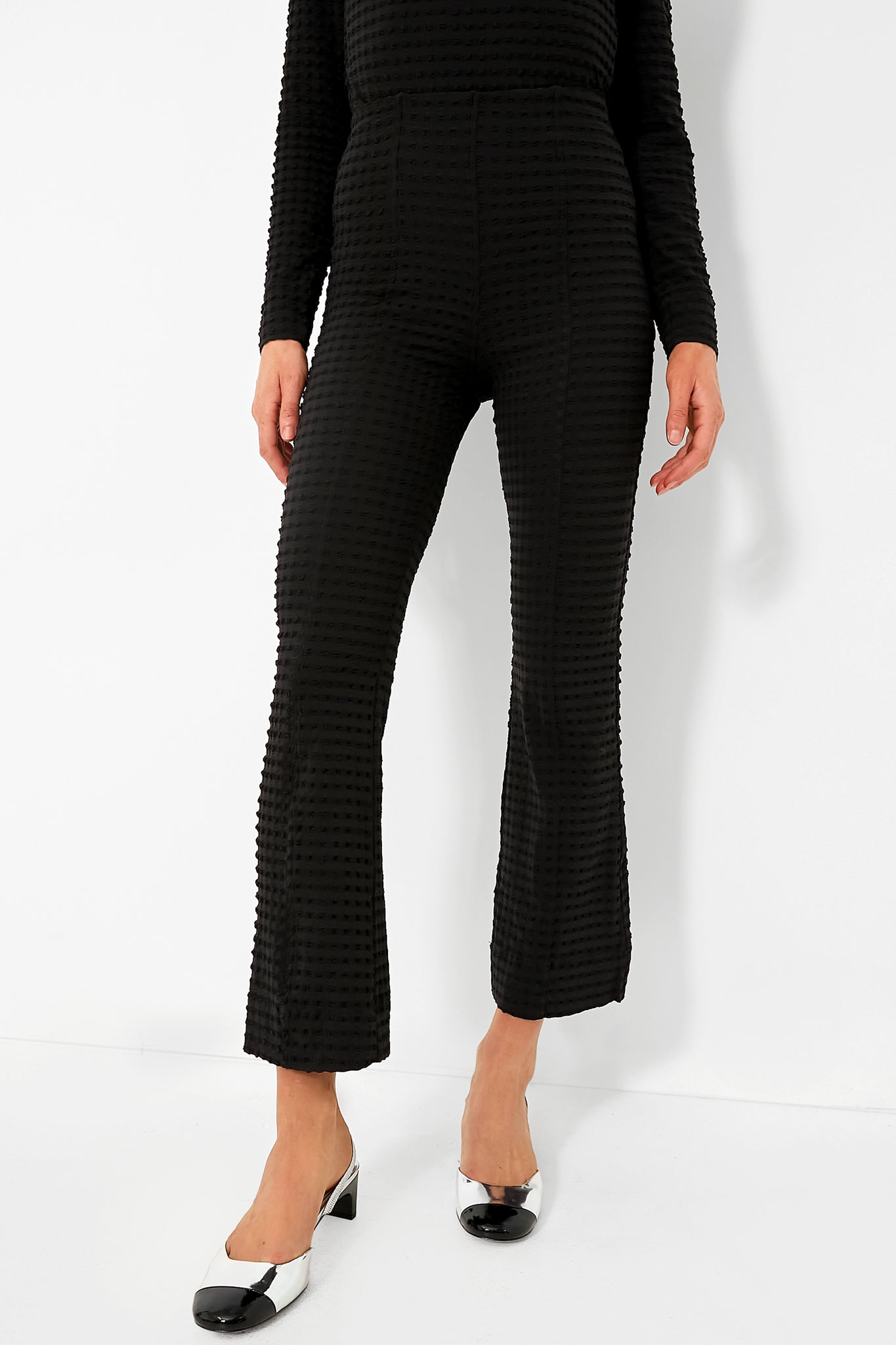 A person wears black textured GANNI pants with a matching long-sleeve top, styled with black and white low-heeled shoes.