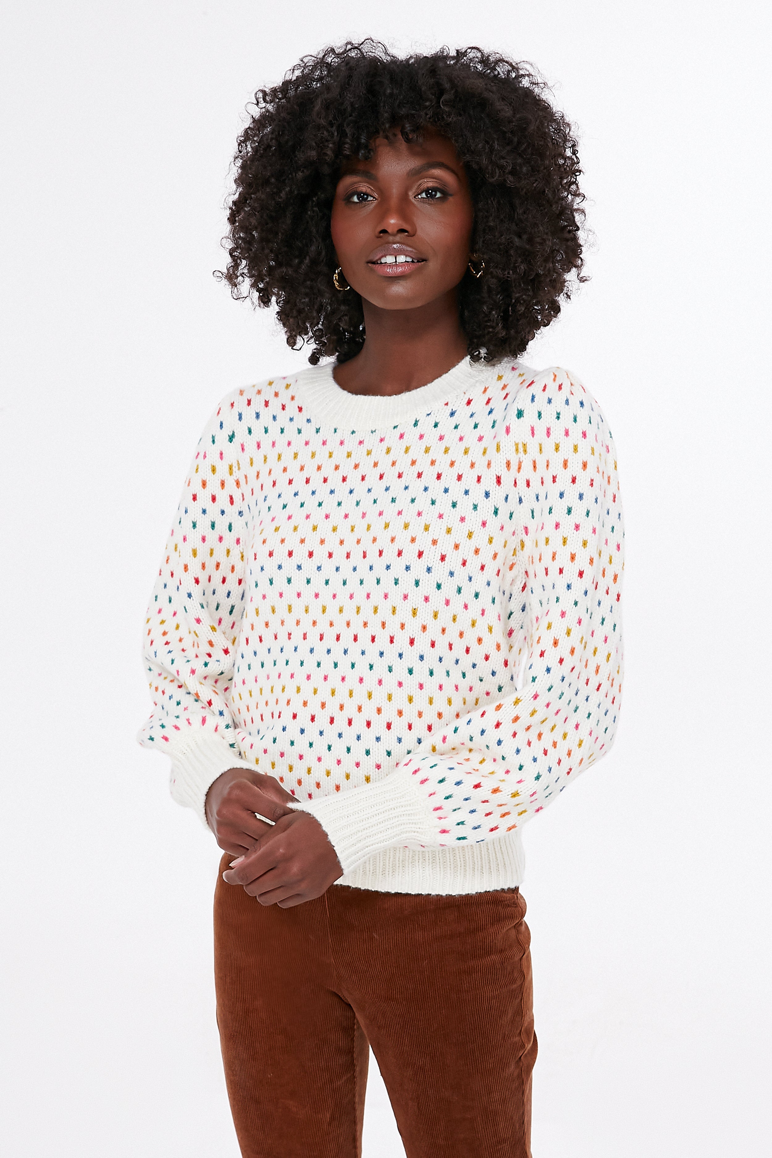 A person wears a Marine Layer eco-friendly sweater featuring a rainbow dot motif and brown pants, hands clasped in front.
