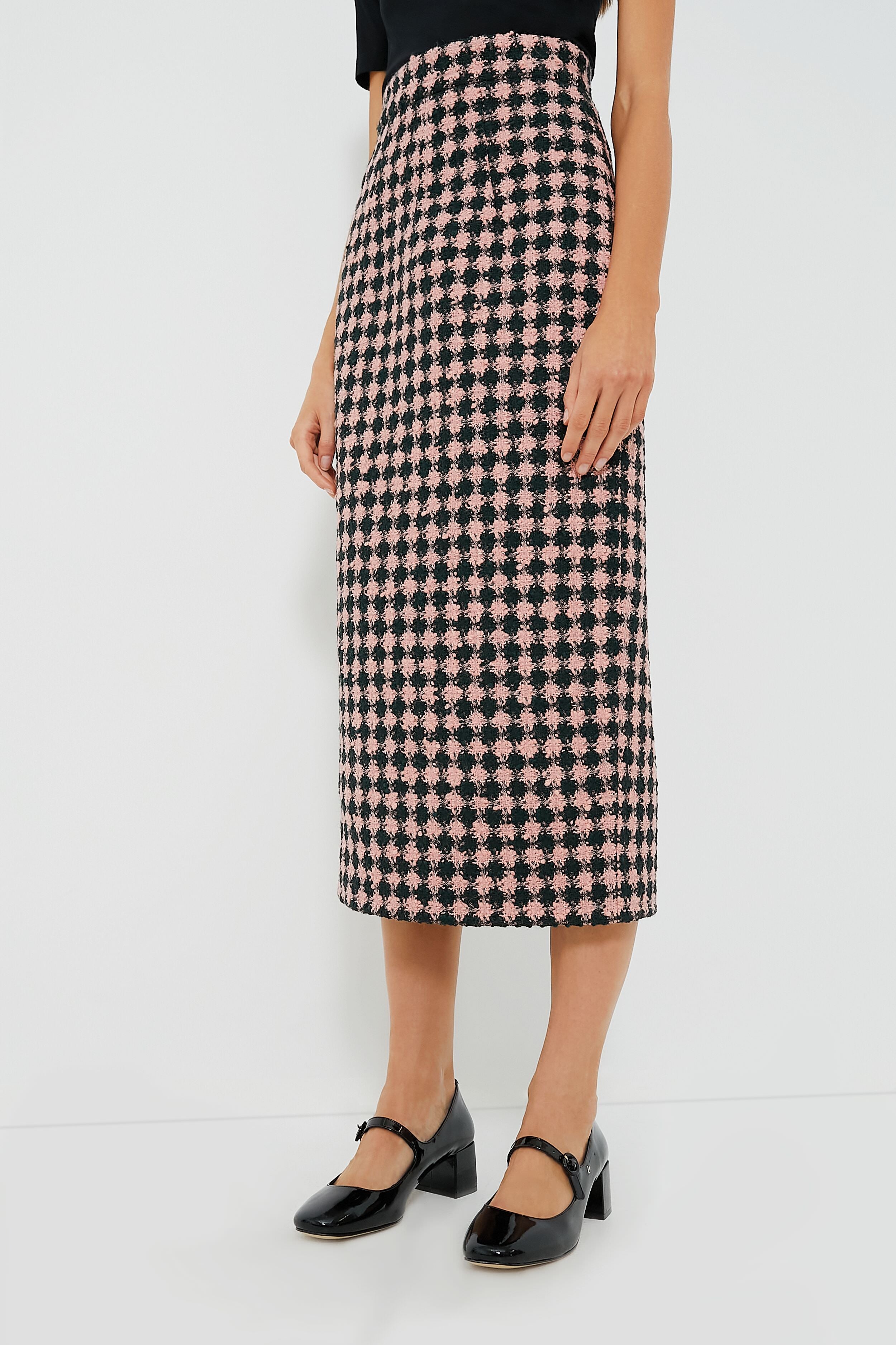 A woman showcases effortless style in an Emilia Wickstead black top and pink-and-black houndstooth skirt, paired with black Mary Jane shoes.