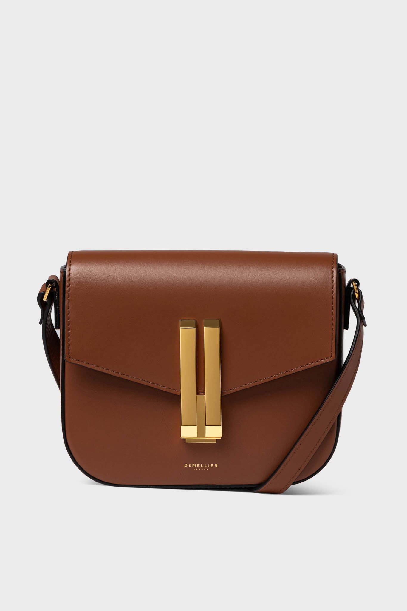 The Demellier brown cowhide leather crossbody bag features a gold rectangular clasp, an adjustable strap, and highlights the quality of handcrafted handbags.